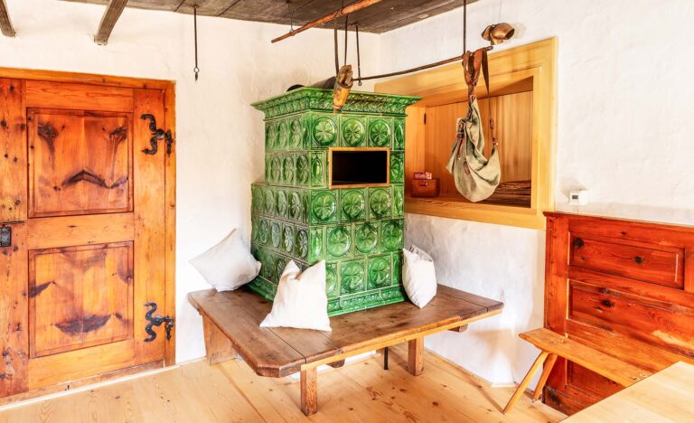 Traditioneller grüner Kachelofen aus Keramik mit Holzbank und weißen Kissen in einem rustikalen Raum mit Holztüren.